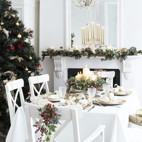 salle à manger blanche décorée pour Noël, table avec vaisselle dorée et blanche, bougies allumées, verres à vin, pièce maîtresse, chaises décorées de baies et de lierre, cheminée en arrière-plan avec miroir sur la cheminée, lustre en or, arbre de Noël avec boules, guirlande de lierre sur la cheminée
