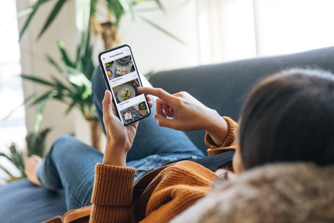 vue d'épaule d'une jeune femme choisissant de la nourriture dans le menu de l'application mobile en position allongée sur le canapé à la maison