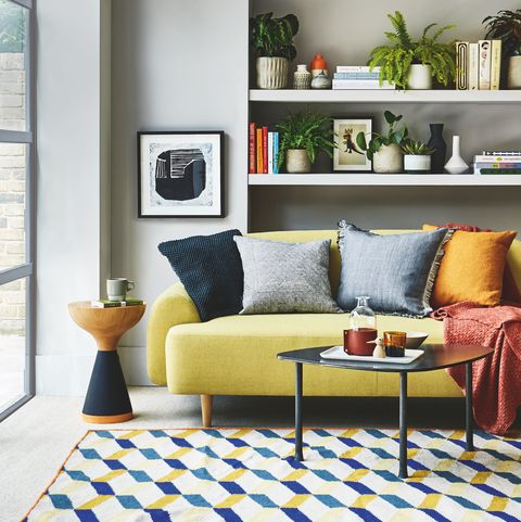 סלון, ספה צהובה מדף לבן מאחור עם שטיח בדוגמת כחול וצהוב על הרצפה