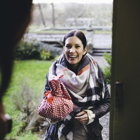 une femme sourit alors qu'elle se tient sur le pas de la porte pour rendre visite à un ami, elle est enveloppée dans des vêtements chauds et tient un cadeau enveloppé dans un tissu à carreaux rouge et blanc