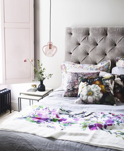 חדר שינה מואר ואוורירי עם מסגרת מיטה מרופדת אפורה וכריות פרחוניות ותריסי חלונות ורודים