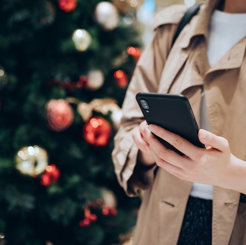 κομμένο πλάνο και μεσαίο τμήμα γυναίκας που χρησιμοποιεί smartphone μπροστά από ένα πολύχρωμο χριστουγεννιάτικο δέντρο την εορταστική περίοδο των Χριστουγέννων